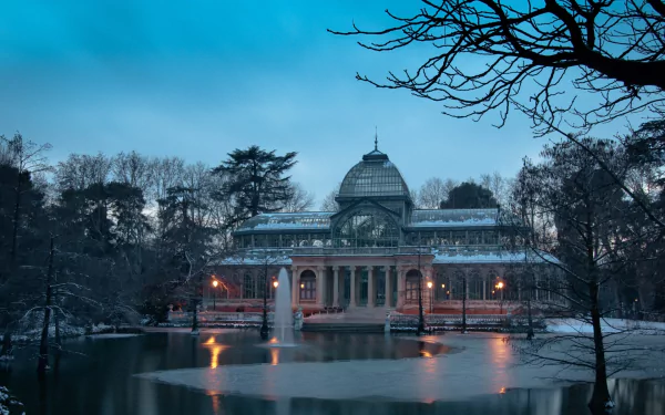 HD desktop wallpaper featuring the illuminated Palacio de Cristal, a man-made glass structure surrounded by trees and a calm reflective pond at dusk.
