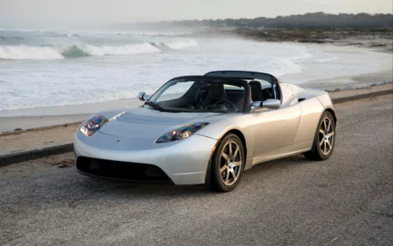 HD wallpaper of a silver Tesla Roadster parked by the beach, suitable as a desktop background.