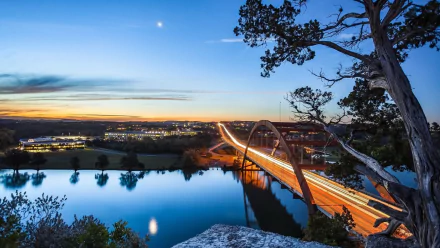 Pennybacker Bridge in Austin, Texas at dusk: illuminated man-made arch and streaking vehicle lights reflected on the water, framed by trees — HD PC desktop wallpaper / background.