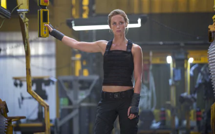 HD wallpaper featuring Emily Blunt in a dynamic pose from the movie Edge of Tomorrow, set in a futuristic industrial environment.