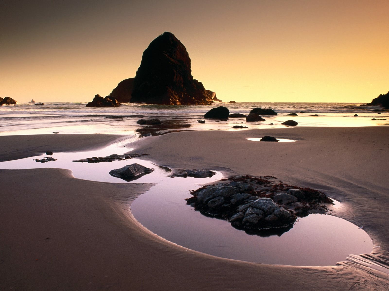 HD desktop wallpaper of a tranquil beach at sunset, with ocean waves gently lapping around dark rocks under a golden sky.