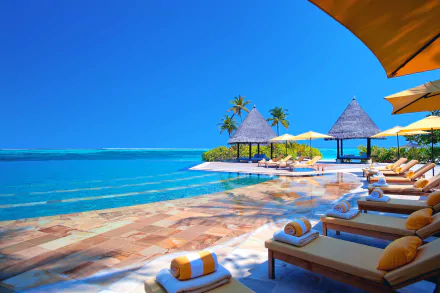 HD wallpaper showing a serene Maldives beach with a luxury swimming pool, bungalows, and lounge chairs under yellow umbrellas, framed by the azure ocean and a clear blue sky.