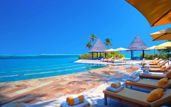 HD wallpaper showing a serene Maldives beach with a luxury swimming pool, bungalows, and lounge chairs under yellow umbrellas, framed by the azure ocean and a clear blue sky.