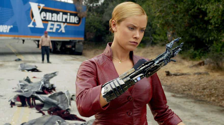 A scene from Terminator 3: Rise of the Machines, featuring a female character examining her robotic arm, set against a backdrop of a truck and scattered debris. HD wallpaper design.