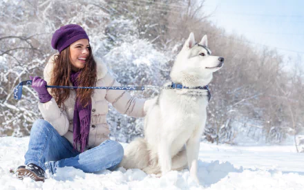 A woman in a purple hat smiles while sitting in the snow with her husky dog, creating a joyful winter scene. This HD desktop wallpaper captures the mood of the season beautifully.