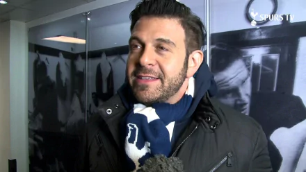 HD wallpaper of a man in a jacket and scarf, smiling during an interview, with a blurred background of photos.