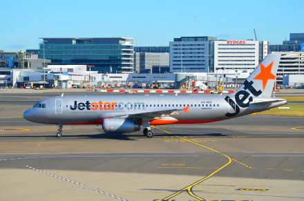 An Airbus A320 Jetstar aircraft at Sydney airport, captured in vibrant detail, showcasing the busy aviation environment and modern airport buildings in the background.