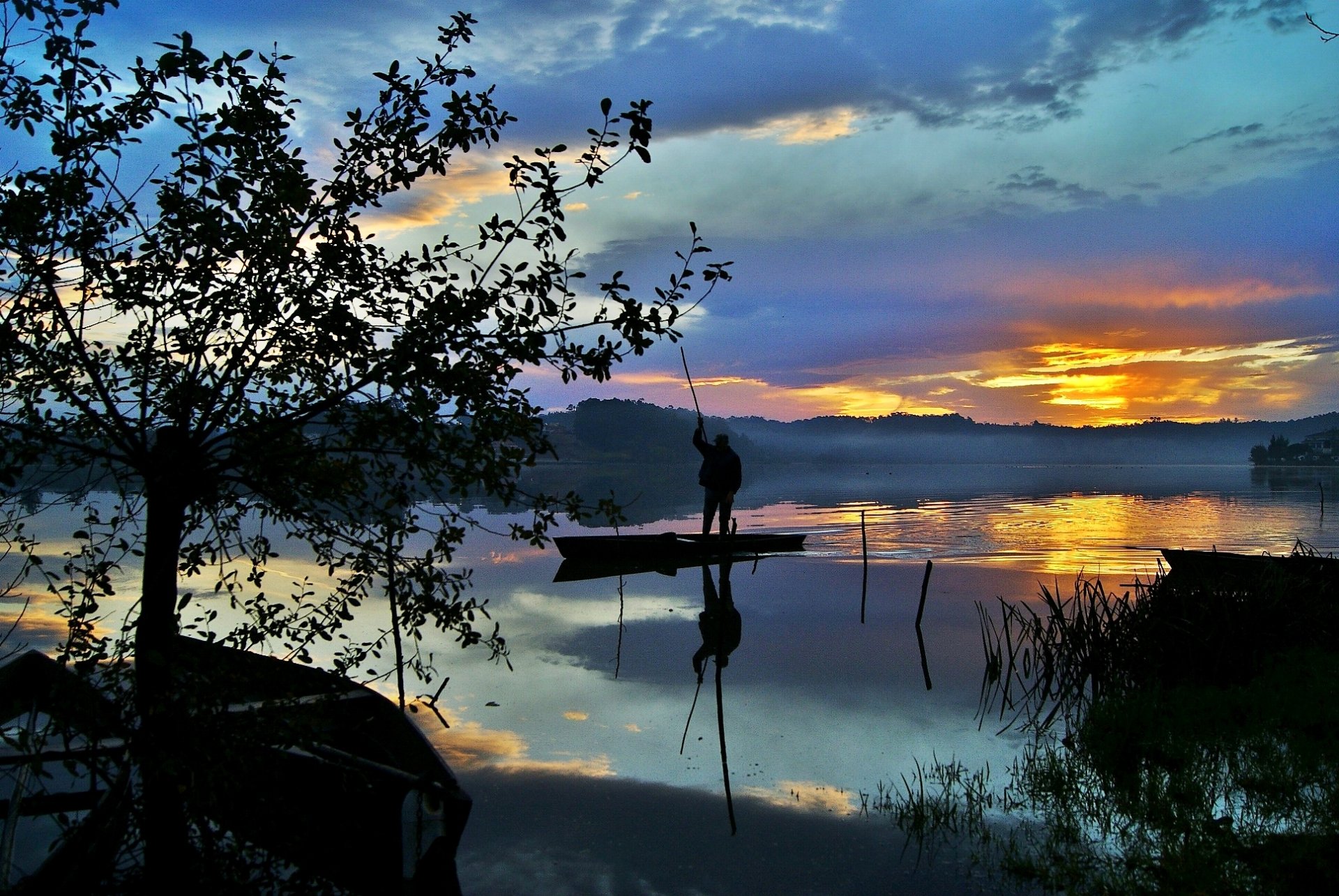 HD photography desktop wallpaper showcasing a serene lake at sunset with vivid sky colors reflected on calm water, framed by silhouetted trees and a boat with a person fishing.