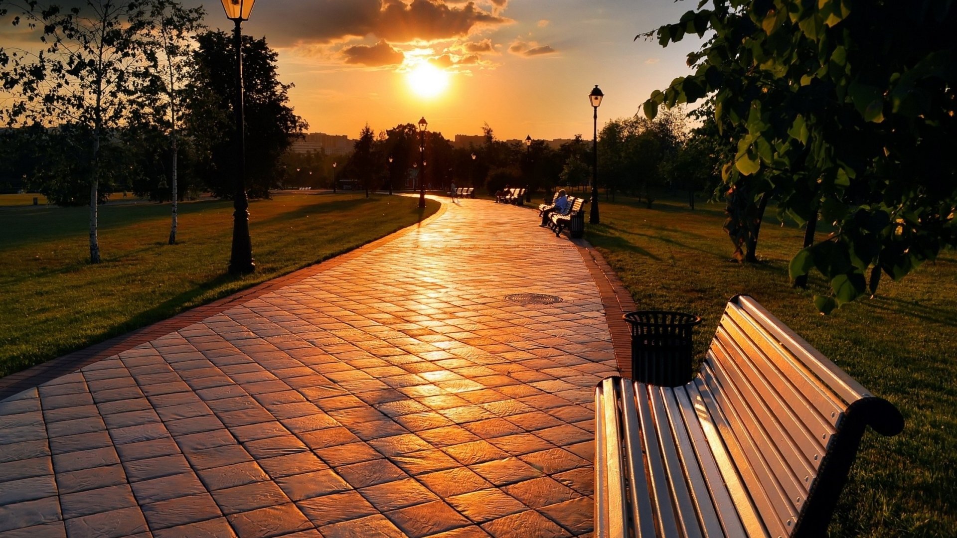 Sunset casting golden light over a park pathway with benches and lampposts, captured in an HD photograph for a peaceful PC desktop wallpaper background.