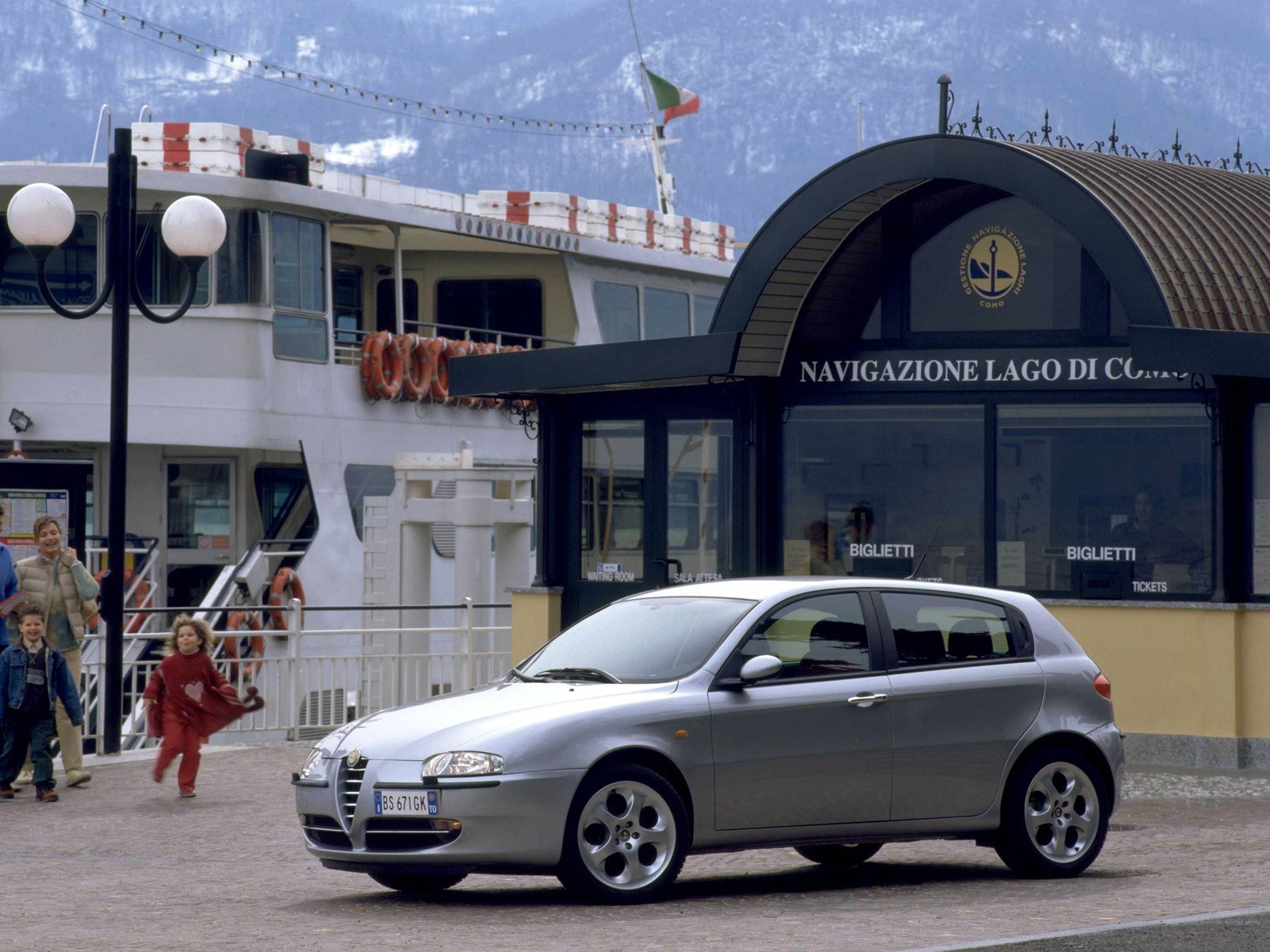 HD PC desktop wallpaper of a silver Alfa Romeo 147 hatchback parked at a lakeside dock, with a ferry and terminal building in the background.