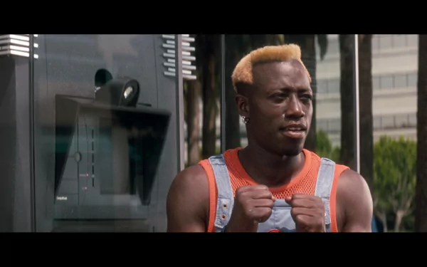 HD PC desktop wallpaper — still from the movie Demolition Man: a man with short bleached hair in an orange tank top and denim overalls, fists clenched and tense in an outdoor urban setting.