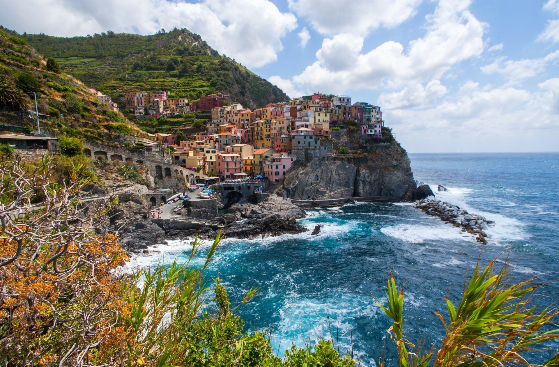 Vibrant coastal village of Manarola in Italy's Liguria region, part of Cinque Terre, perched on cliffs overlooking the blue Mediterranean Sea under a partly cloudy sky.