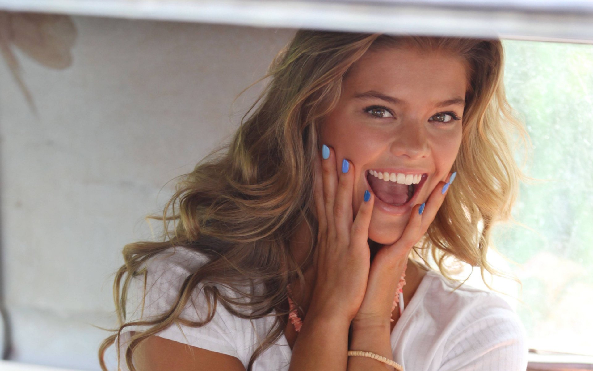Sunlit portrait of a smiling blonde woman with long wavy hair and blue nail polish, hands framing her face — 2K Quad HD desktop wallpaper.