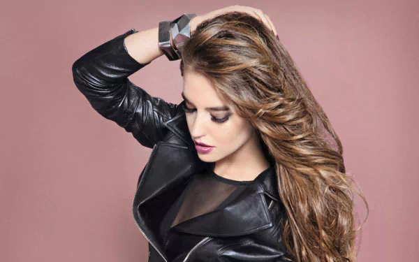 HD desktop wallpaper featuring Spanish woman Clara Alonso with long flowing hair, wearing a black leather jacket against a muted pink background.