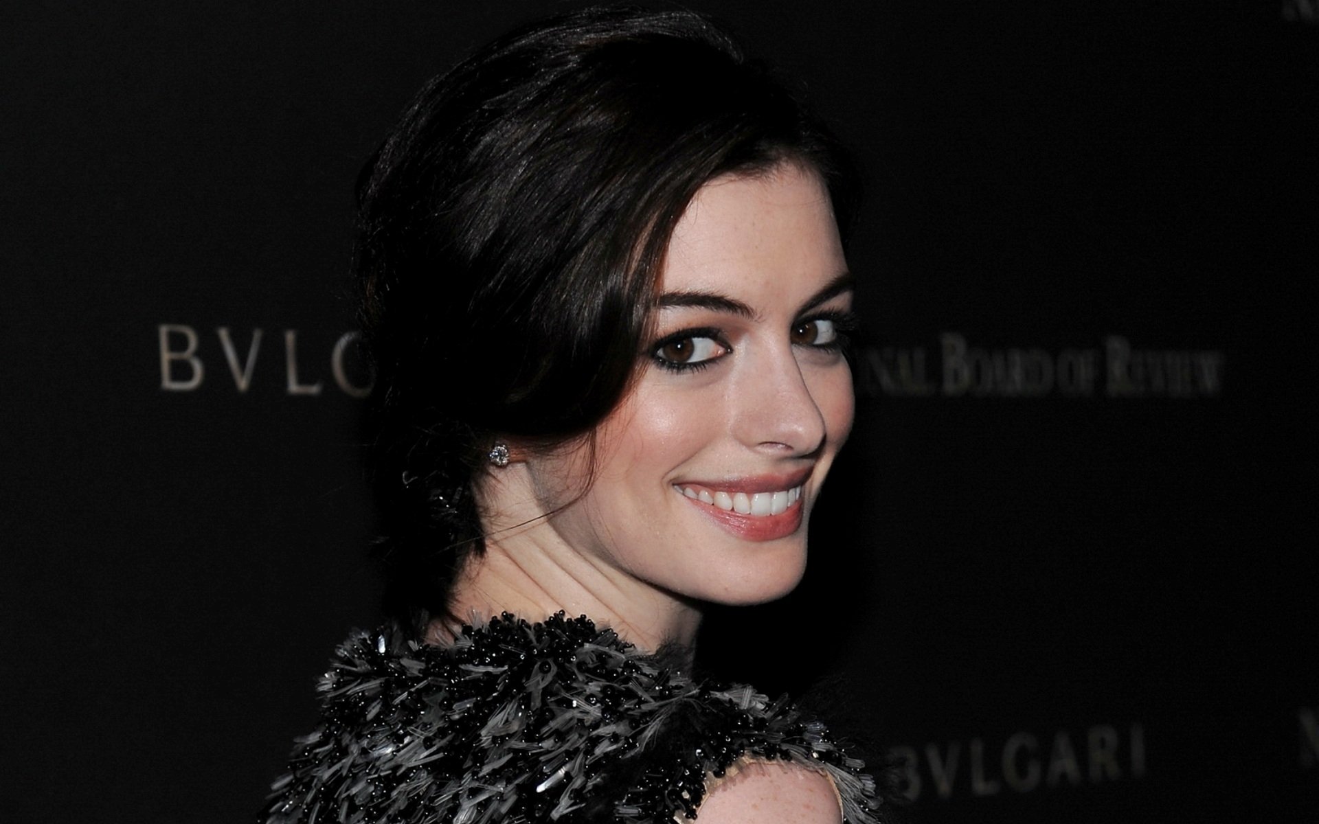 HD PC desktop wallpaper featuring celebrity Anne Hathaway smiling over her shoulder against a dark background with subtle brand text.