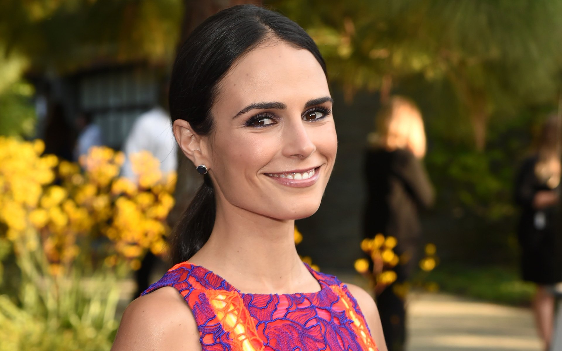 HD desktop wallpaper featuring Brazilian actress and celebrity Jordana Brewster smiling outdoors with blurred greenery and yellow flowers in the background.