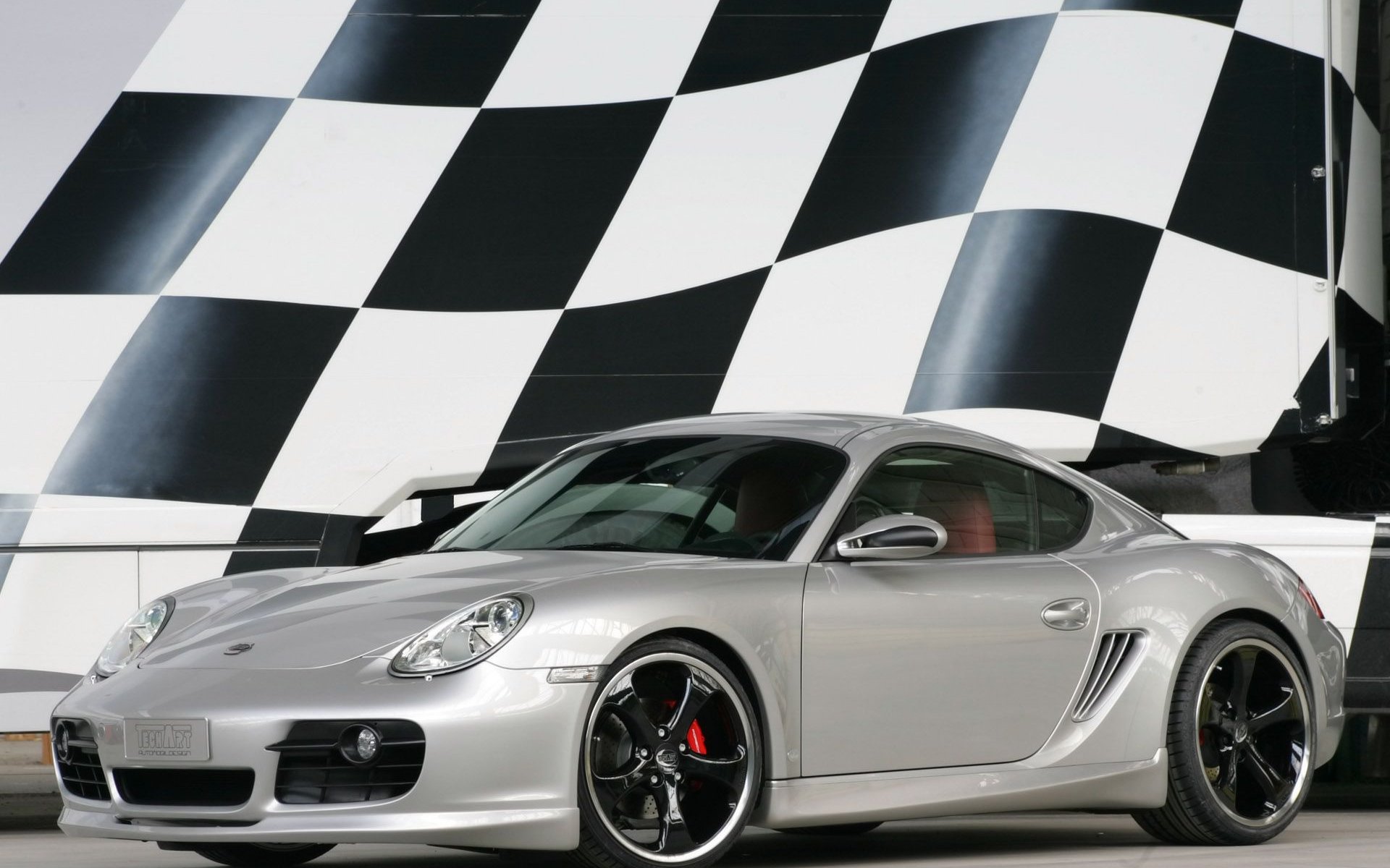 Silver Porsche Cayman sports car parked indoors with a black-and-white checkered wall in the background, presented as an HD PC desktop wallpaper and background.