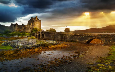 HD desktop wallpaper featuring the man-made Eilean Donan Castle under a dramatic sky with sunlight breaking through clouds, highlighting the historic stone bridge and surroundings.
