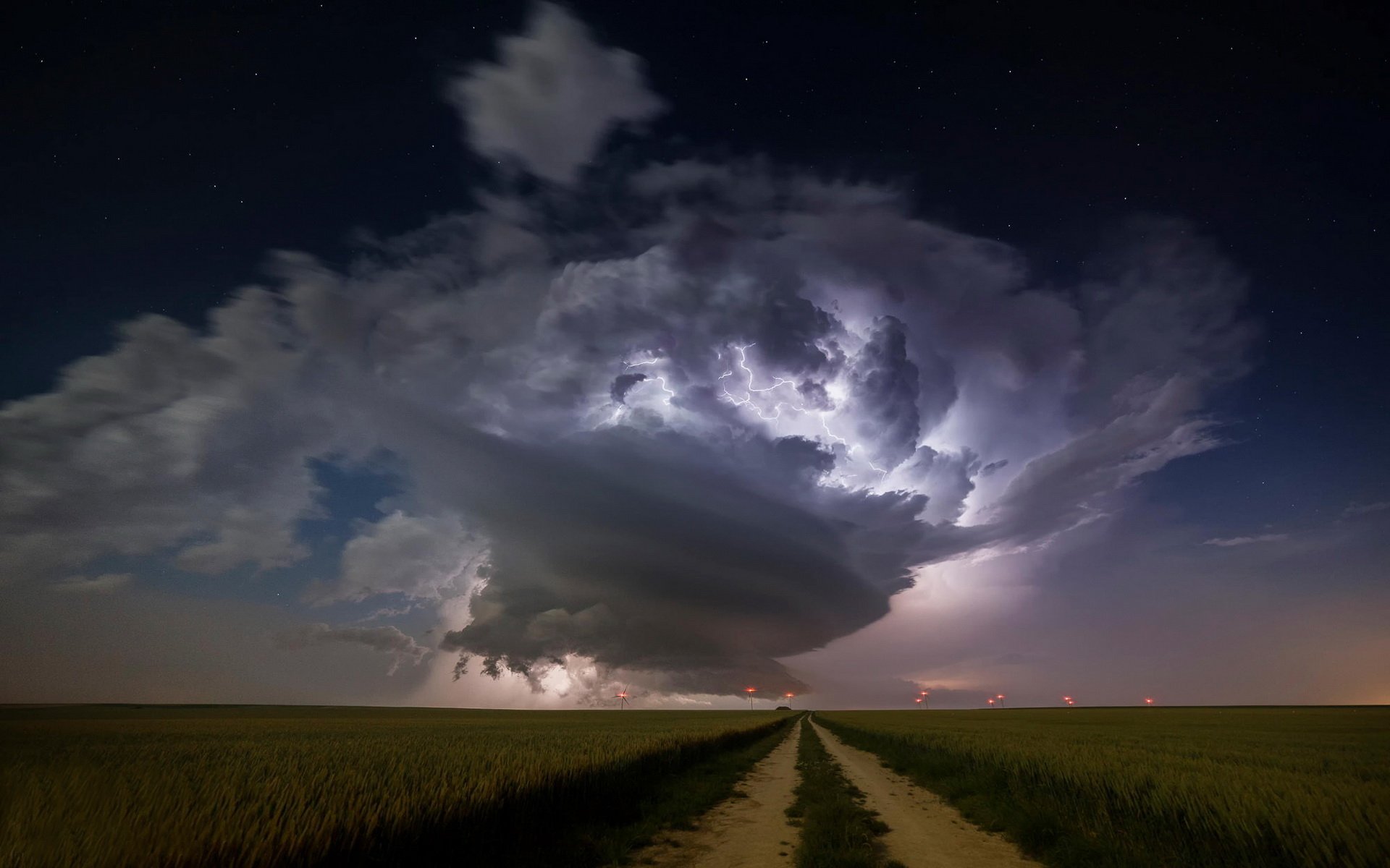 HD PC desktop wallpaper and background: dramatic nighttime nature storm — a massive illuminated supercell thundercloud with lightning over flat plains, a dirt road leading toward it.
