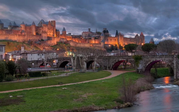 A stunning HD wallpaper of Carcassonne, showcasing its illuminated medieval fortress against a dramatic sky, with scenic landscapes and a flowing river in the foreground.