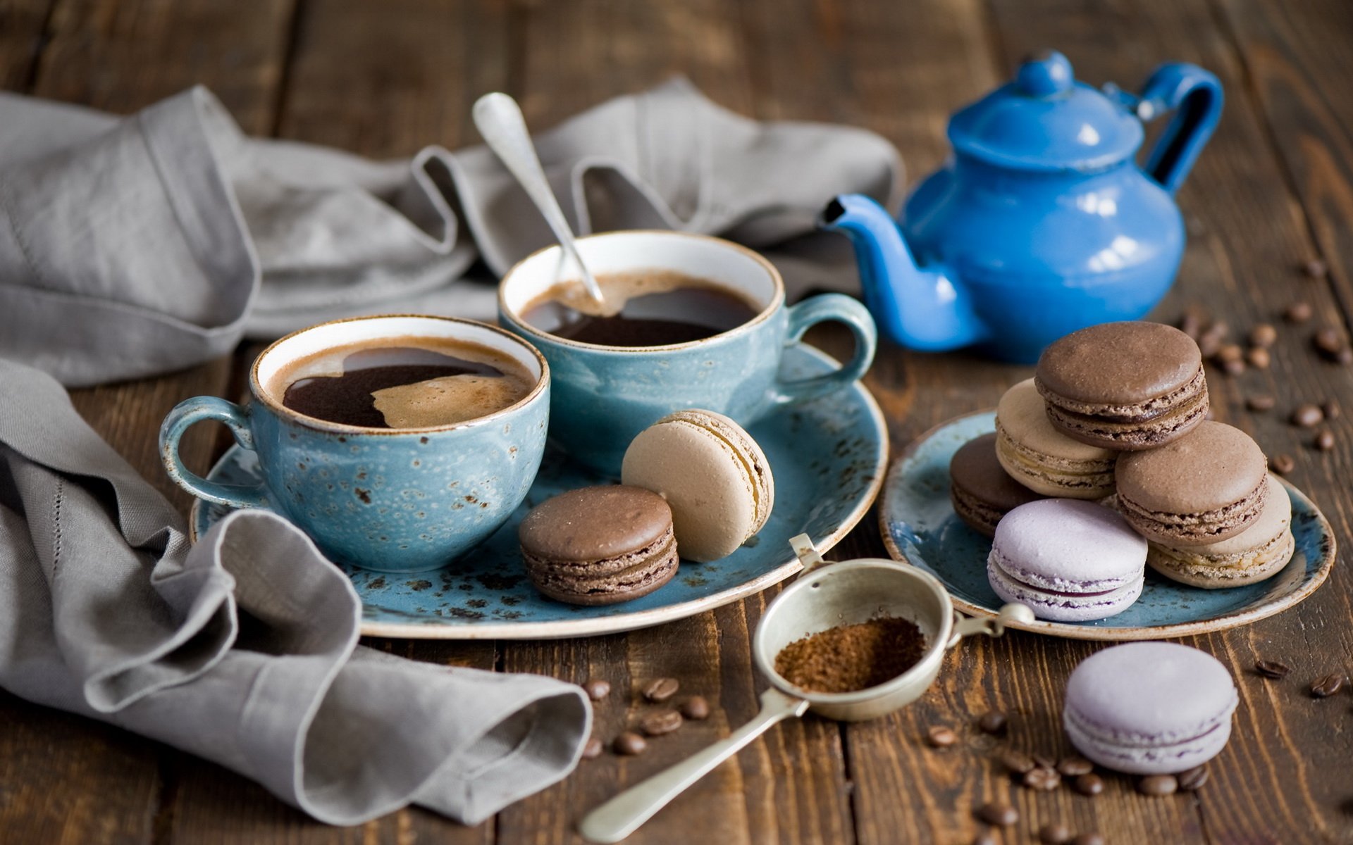 HD PC desktop wallpaper showing two cups of coffee with macarons on rustic wooden table, accompanied by a blue teapot and coffee beans scattered around.