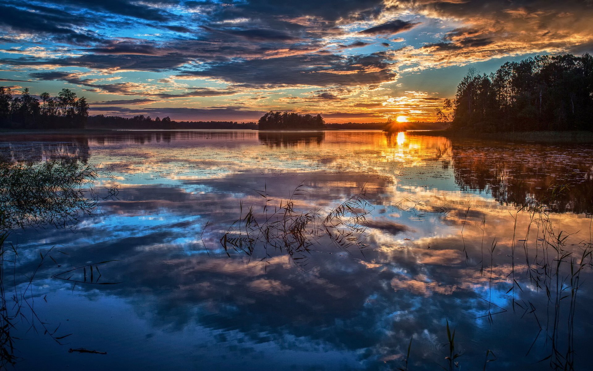 HD PC desktop wallpaper: serene sunset over a lake with dramatic clouds and golden-orange sky mirrored in glassy water, trees silhouetted along the horizon.