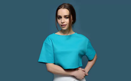 A stunning HD wallpaper featuring brunette actress Emilia Clarke, posing confidently in a blue crop top against a teal background.