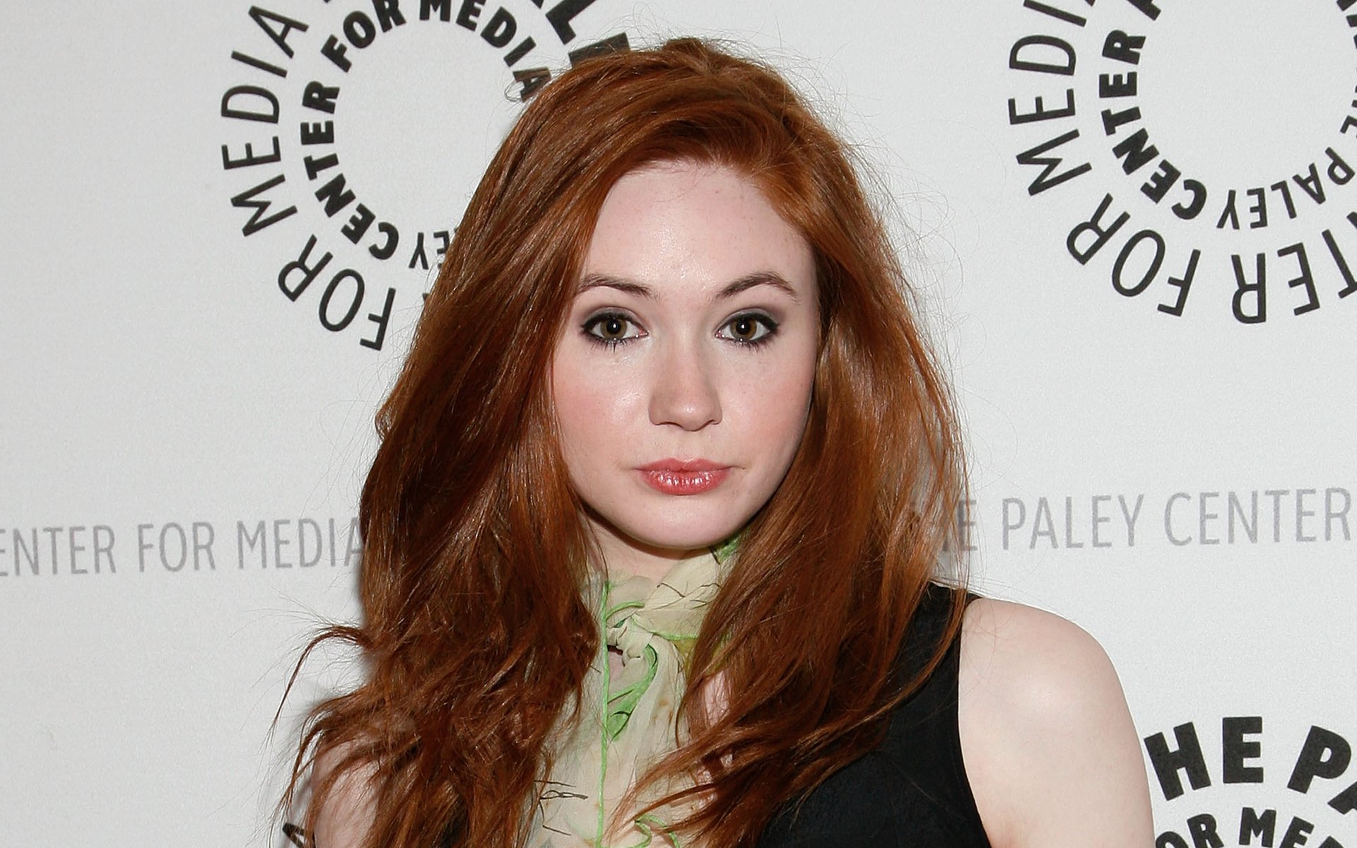 HD desktop wallpaper close-up of a red‑haired woman in a black dress and green scarf posing before a media step-and-repeat backdrop.