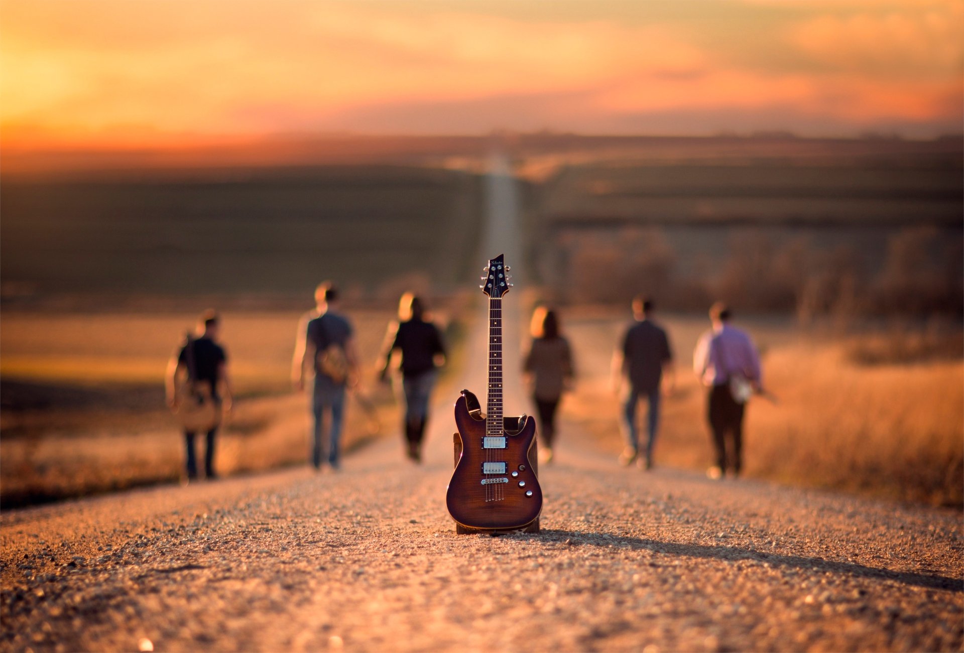 Depth of field image of five people walking down a road with a guitar at the center, set against a scenic sunset. HD wallpaper.