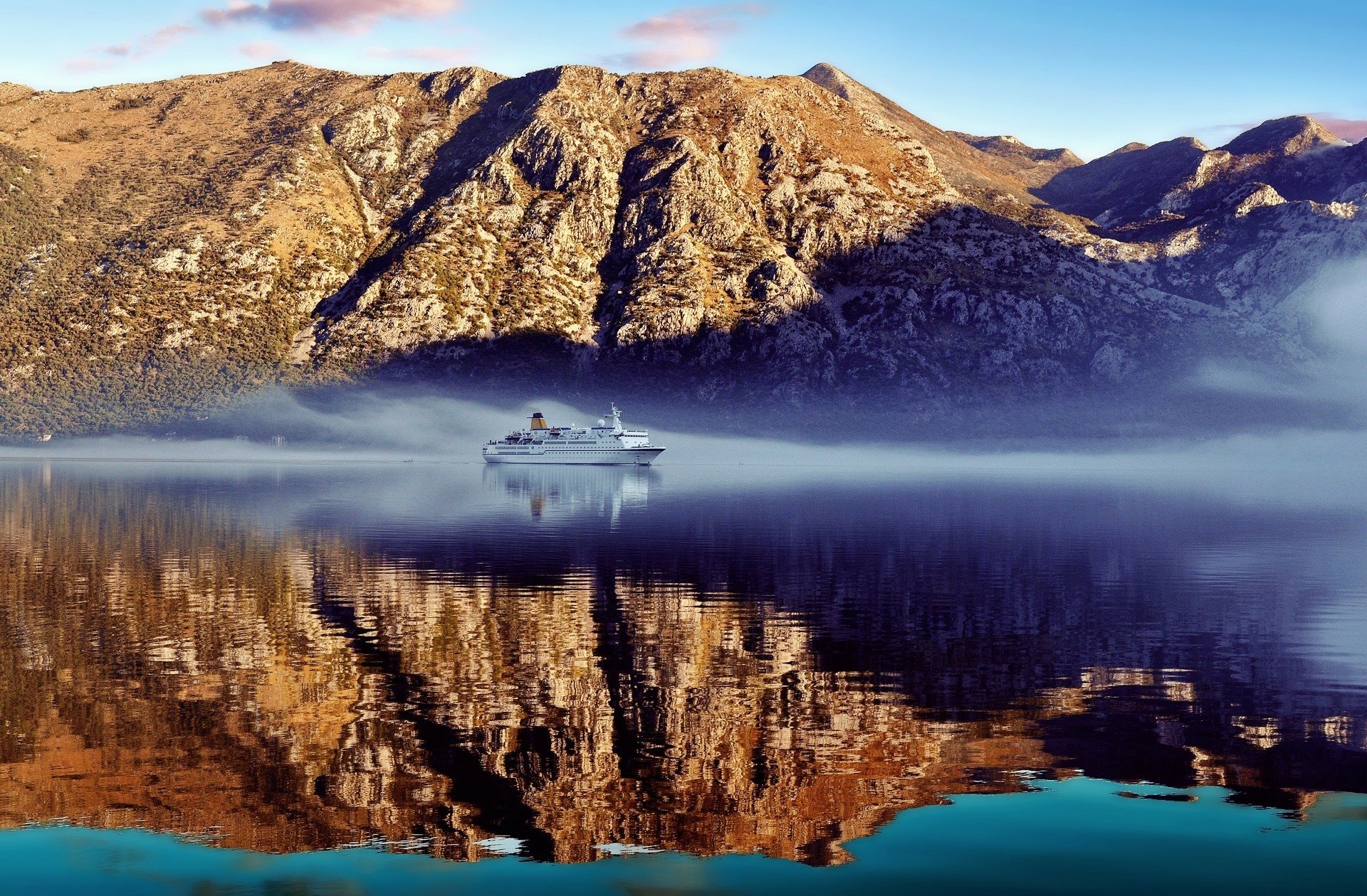 A serene fjord scene with a liner sailing through calm waters, perfectly reflecting the rugged mountain landscape under clear blue skies. HD desktop wallpaper.