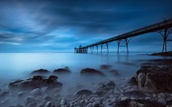 HD PC desktop wallpaper showing a foggy seaside scene with pebbles in the foreground and a long man-made pier extending into the misty sea.