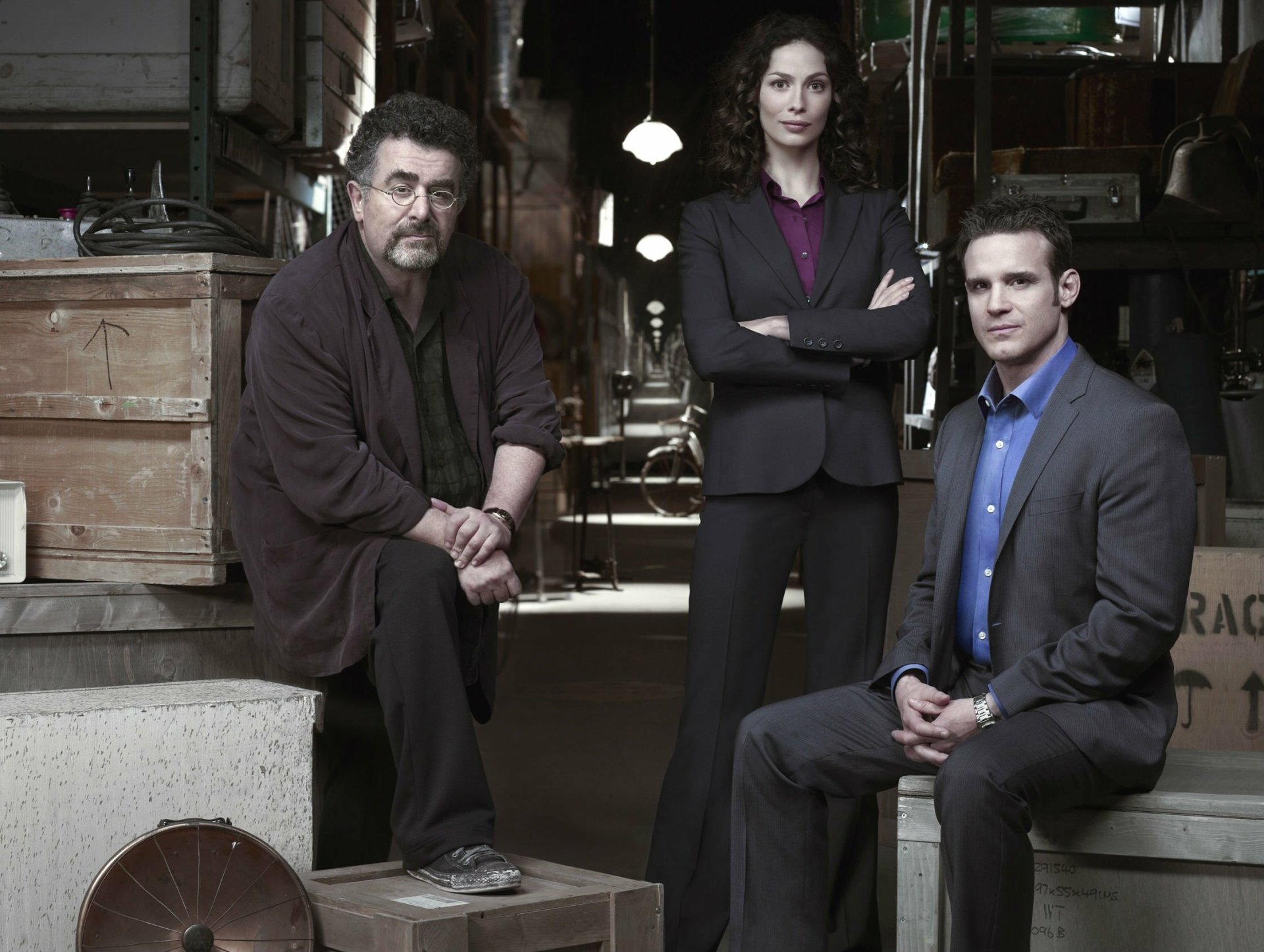 HD PC desktop wallpaper featuring main cast of the TV show Warehouse 13 standing in a dimly lit warehouse filled with crates and mysterious artifacts.
