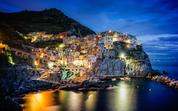 HD desktop wallpaper showing the man-made colorful cliffside village of Manarola illuminated at night against a deep blue sky and calm coastal waters.