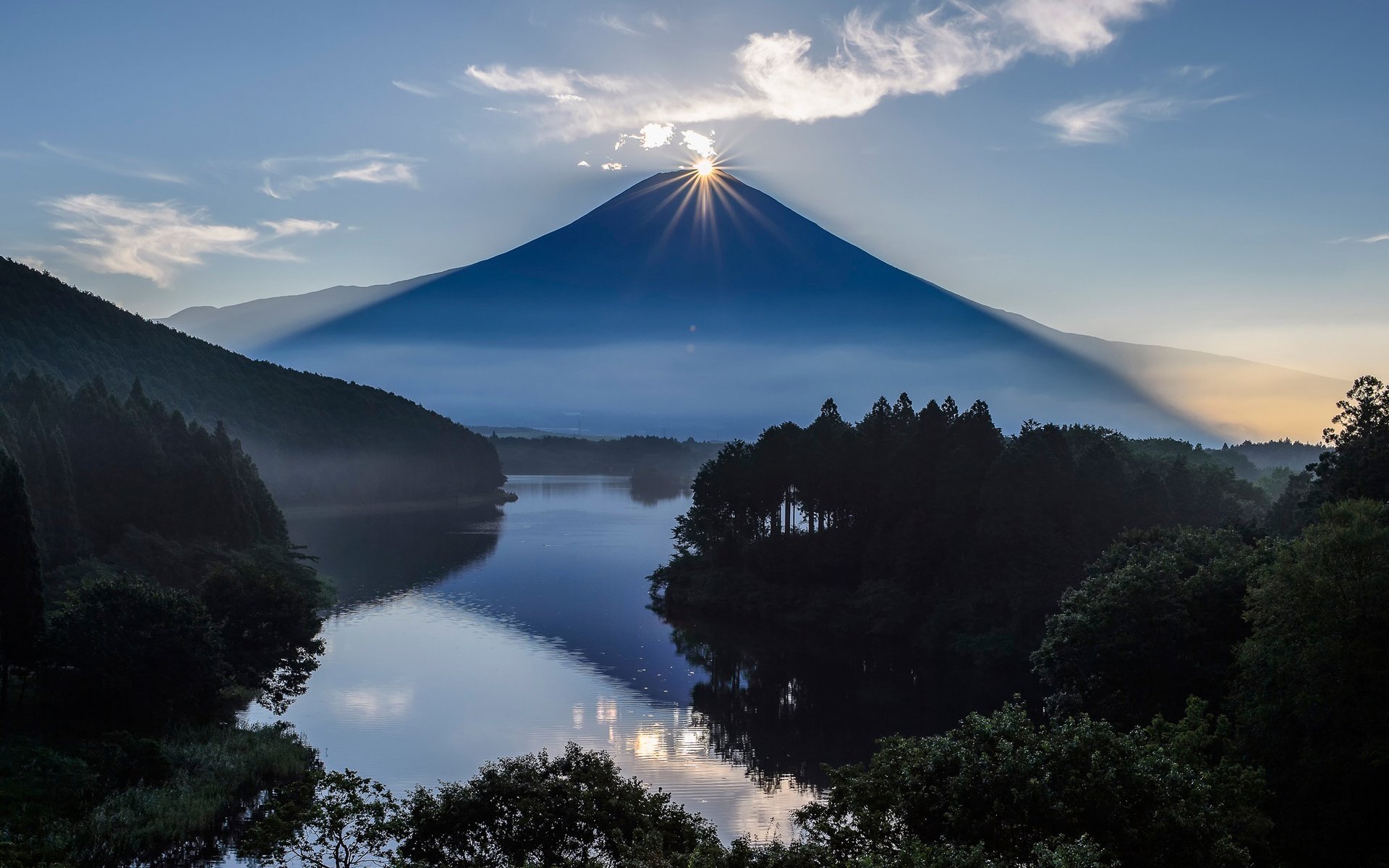 HD PC desktop wallpaper: volcanic mountain at sunrise with a starburst over the summit, mirrored in a calm lake and framed by dark, forested shores.