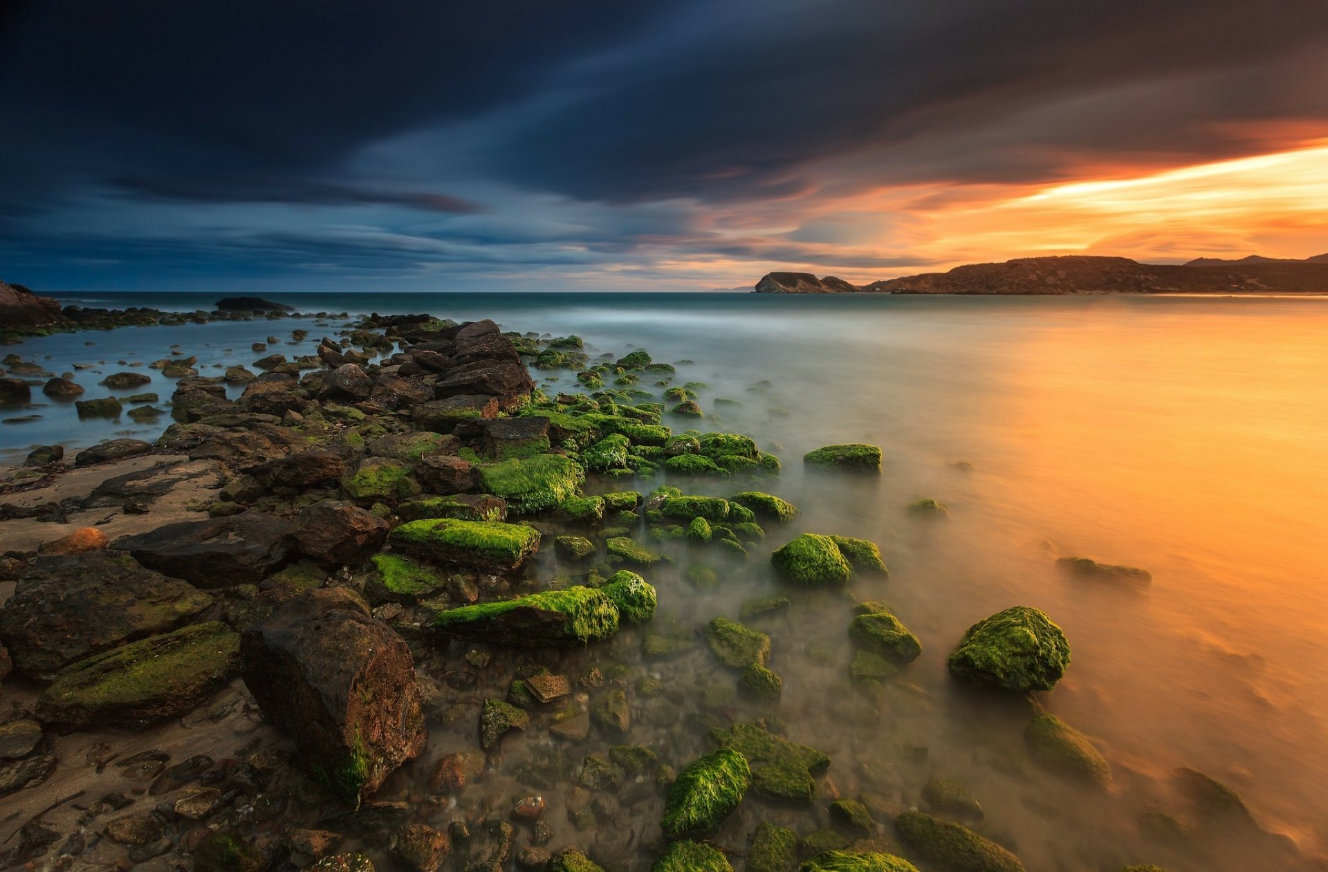 HD PC desktop wallpaper: seashore at sunset with a golden-orange sky and dark clouds, smooth reflective water and moss-covered rocks leading toward distant headlands.