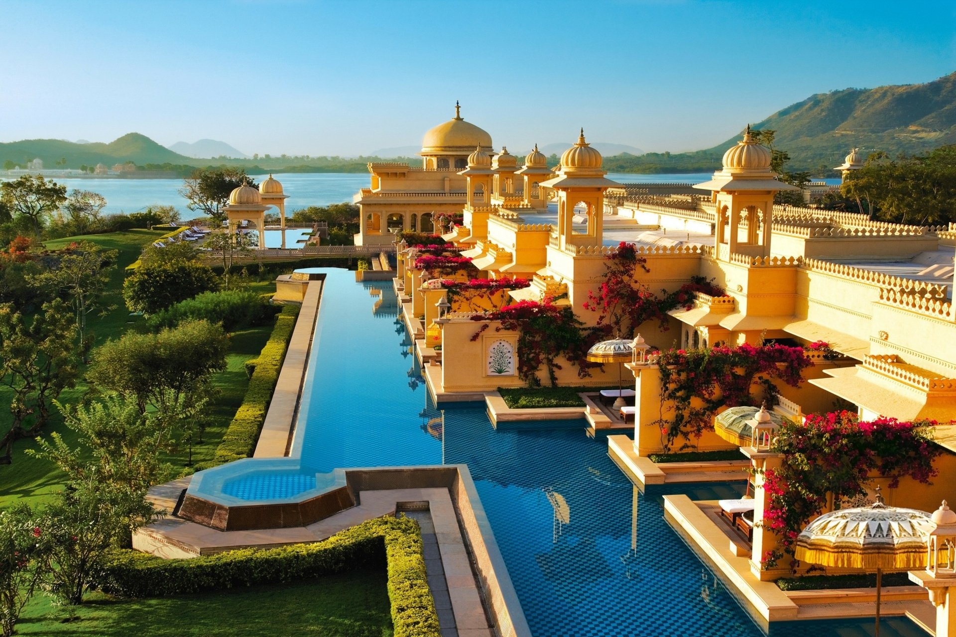 A stunning HD desktop wallpaper of Oberoi Udaivilas hotel in Udaipur, Rajasthan, India, showcasing luxurious architecture by the water with scenic hills in the background.