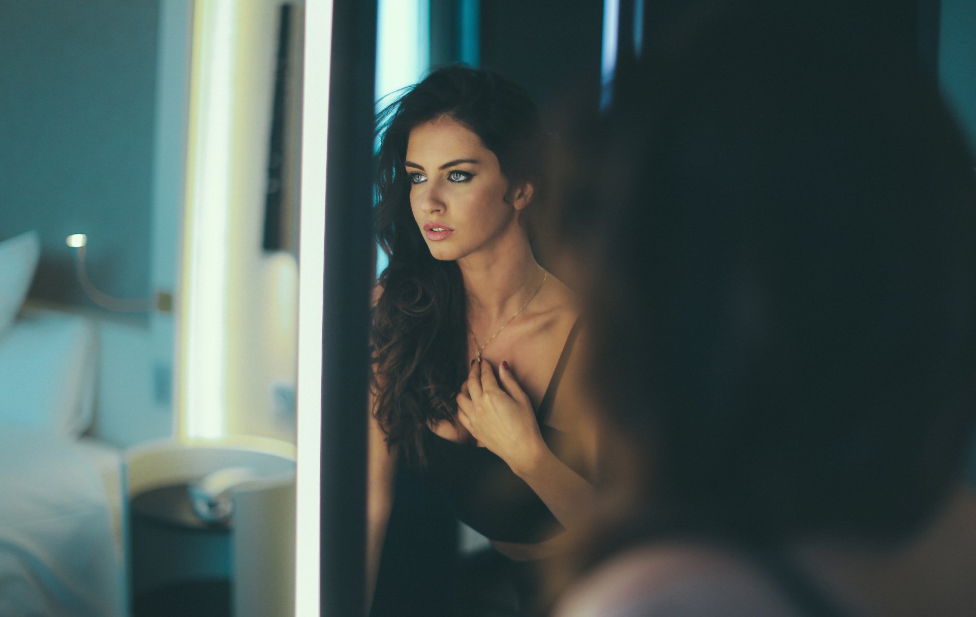 A brunette woman with striking blue eyes gazes thoughtfully into a mirror, capturing a moment of elegance and beauty in a stylish, modern setting. High-definition wallpaper quality.