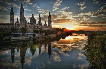 A stunning sunset over the river in Zaragoza, Spain, with the Basilica of Our Lady of the Pillar reflected in the water, capturing a serene moment of beauty and architecture.