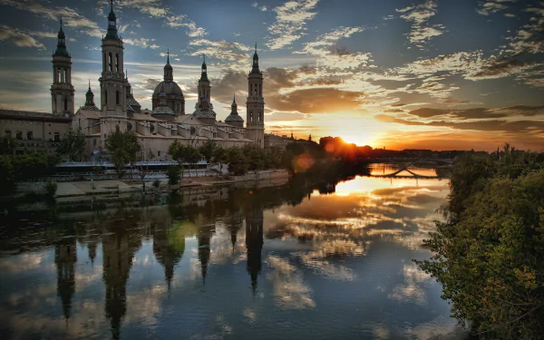 A stunning sunset over the river in Zaragoza, Spain, with the Basilica of Our Lady of the Pillar reflected in the water, capturing a serene moment of beauty and architecture.