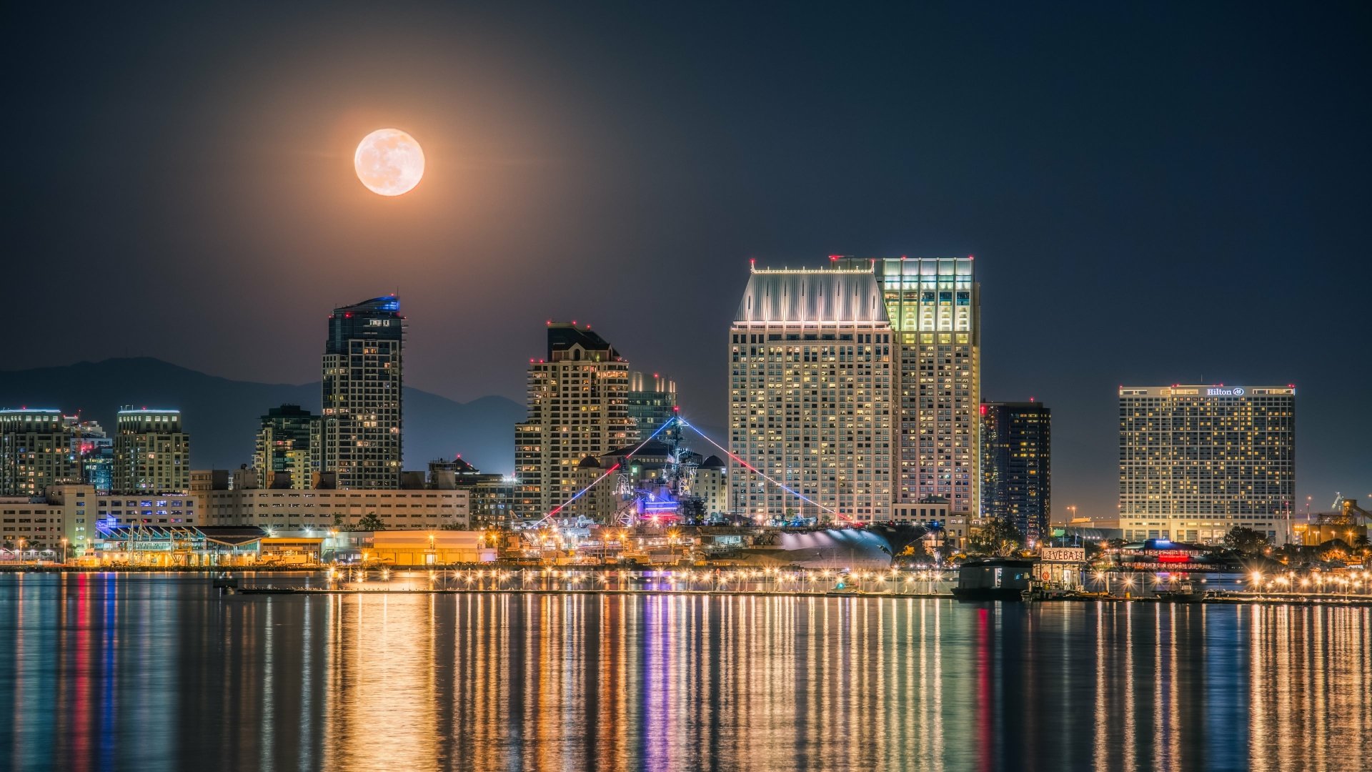 4K Ultra HD PC desktop wallpaper of San Diego, California harbor at night: moonlit sky over illuminated skyscrapers and waterfront buildings, a docked ship and man-made skyline reflected in calm water.