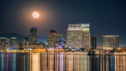 4K Ultra HD PC desktop wallpaper of San Diego, California harbor at night: moonlit sky over illuminated skyscrapers and waterfront buildings, a docked ship and man-made skyline reflected in calm water.