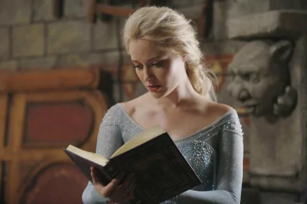 Elsa from Once Upon A Time is depicted reading a book, dressed in her signature blue gown, set against a medieval backdrop, captured in this HD desktop wallpaper.