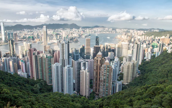 A 4K Ultra HD view of Hong Kong's dense skyline with towering man-made skyscrapers framed by green hills under a partly cloudy sky.