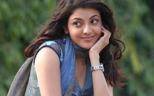 An HD desktop wallpaper featuring Bollywood celebrity Kajal Aggarwal with a serene expression, resting her face on her hand against a backdrop of lush greenery.