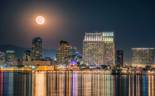 4K Ultra HD PC desktop wallpaper of San Diego, California harbor at night: moonlit sky over illuminated skyscrapers and waterfront buildings, a docked ship and man-made skyline reflected in calm water.