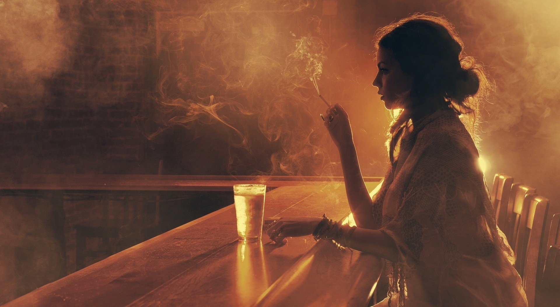 A woman sits contemplatively at a dimly lit bar, holding a cigarette. The rich, moody atmosphere makes this high-definition image a striking desktop wallpaper and background.