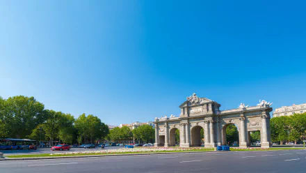 HD desktop wallpaper featuring the man-made Puerta de Alcalá monument under a clear blue sky with surrounding greenery and a quiet street in the foreground.