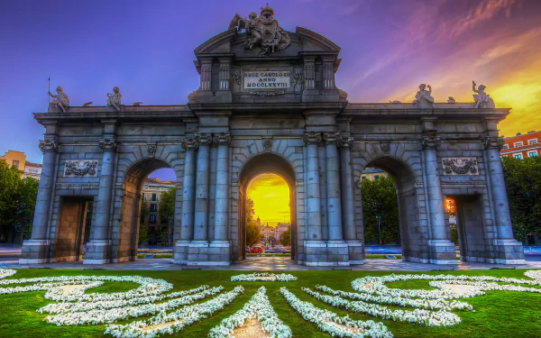HD desktop wallpaper showcasing the man-made Puerta de Alcalá monument at sunset, with vibrant sky colors and floral patterns in the foreground.