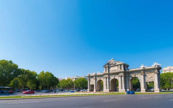 HD desktop wallpaper featuring the man-made Puerta de Alcalá monument under a clear blue sky with surrounding greenery and a quiet street in the foreground.