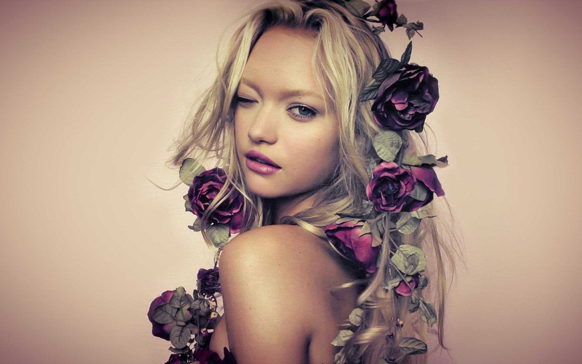 Portrait of blonde Australian model Gemma Ward with blue eyes, soft pink lipstick, and roses entwined in her hair, set against a neutral background.